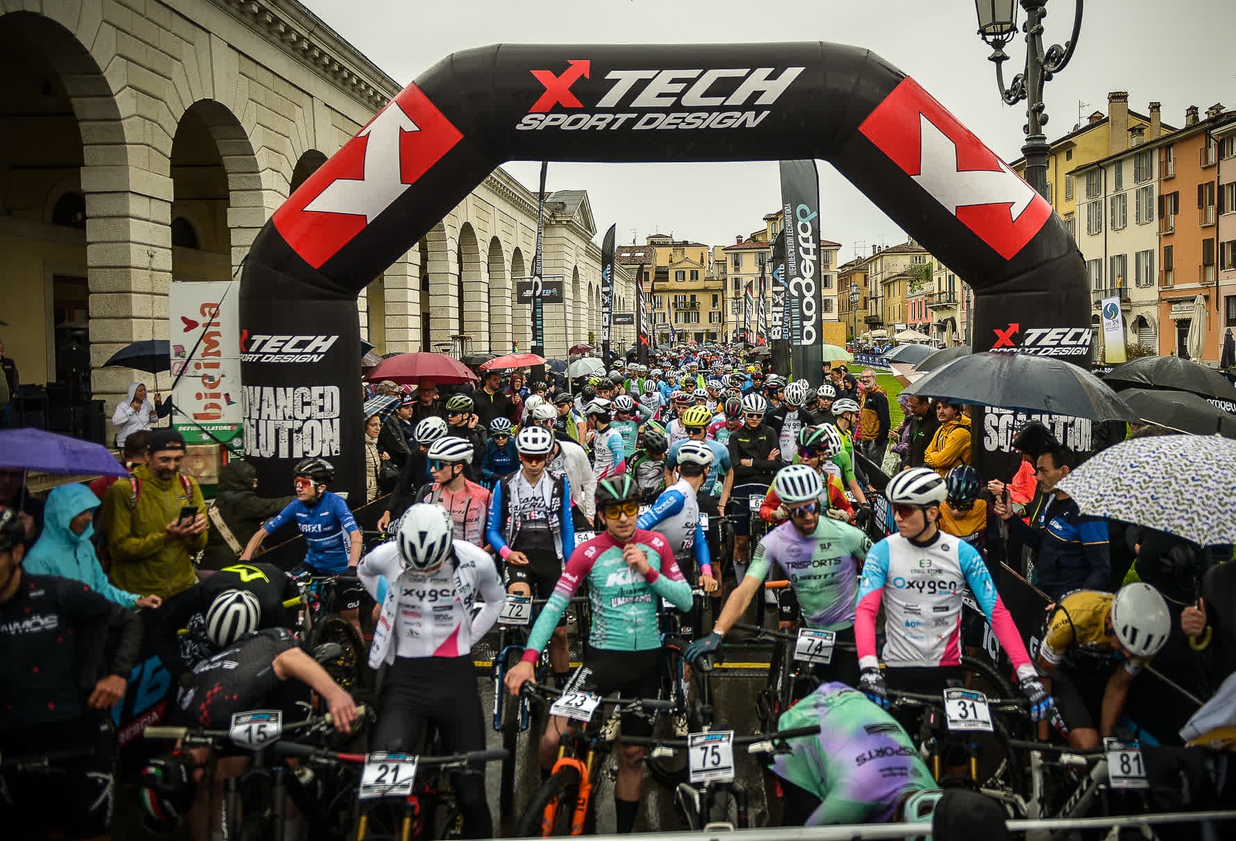 Ciclisti mountain bike radunati sotto l'arco gonfiabile XTech alla partenza della Gran Fondo MTB XTech Città di Brescia in Piazza Arnaldo, sotto la pioggia.